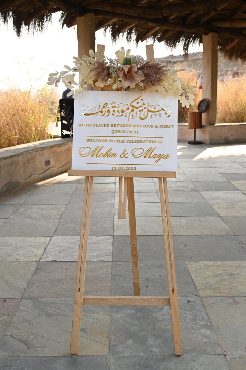 Puede incluir: Un cartel blanco con letras doradas en un caballete. El cartel dice "And He Placed Between You Love & Mercy, Quran 30:21, Welcome to the Celebration of Mobin & Maya, 12.05.2022".