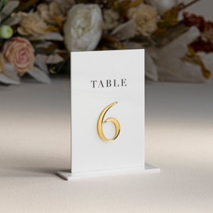 Może przedstawiać: Biała karta numeru stołu z napisem "TABLE" i numerem "6" złotymi literami.