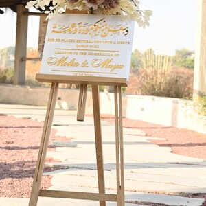 Puede incluir: Un cartel blanco con letras doradas en un caballete. El cartel dice "And He Placed Between You Love & Mercy Quran 90:21 Welcome to the Celebration of Melin & Maya 12.08.2022".