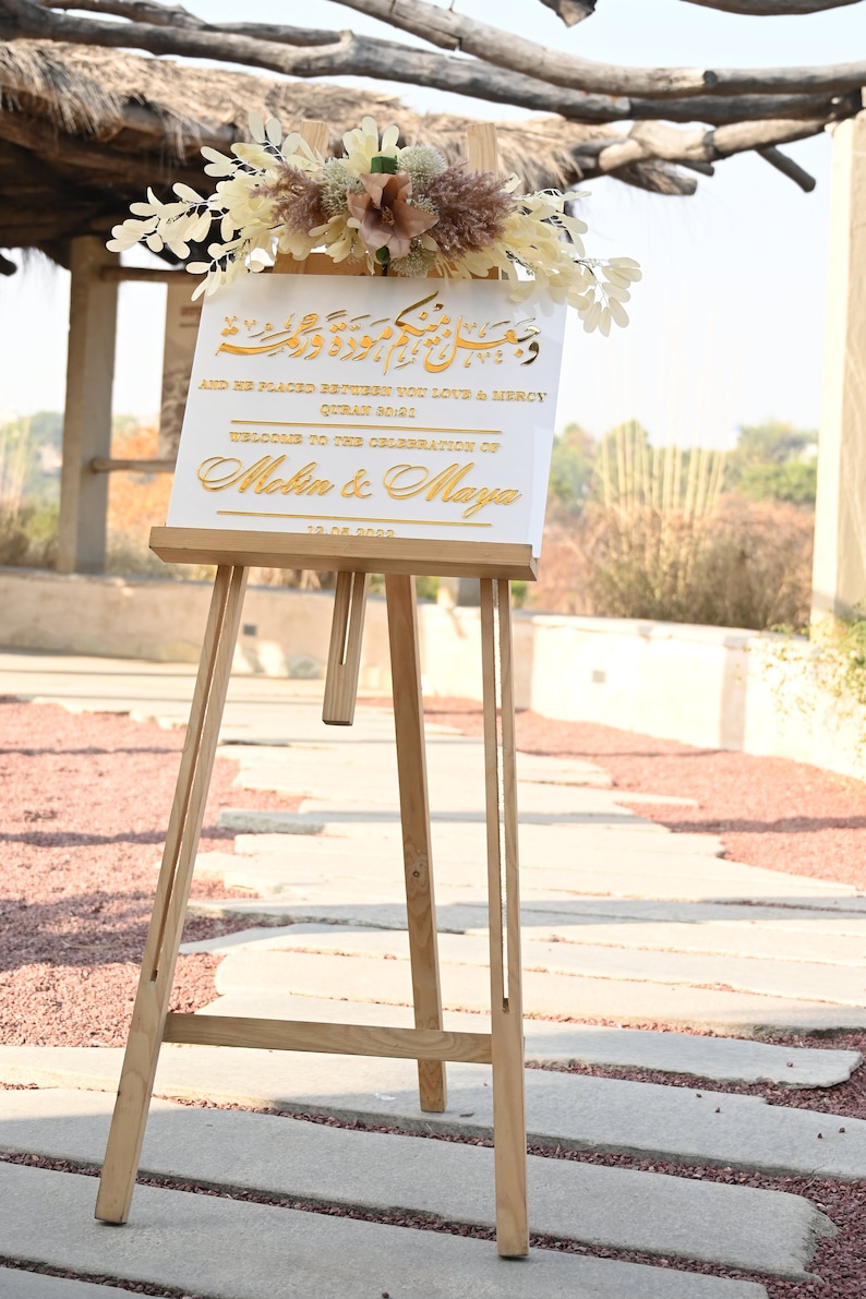Puede incluir: Un cartel blanco con letras doradas en un caballete. El cartel dice "And He Placed Between You Love & Mercy Quran 90:21 Welcome to the Celebration of Melin & Maya 12.08.2022".