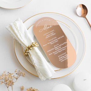 May include: A rose gold wedding menu card with rounded edges, featuring the names "LAUREN & ERICK" and a list of menu items. The card rests on a white plate with a gold rim, alongside a folded white napkin and a gold napkin ring.