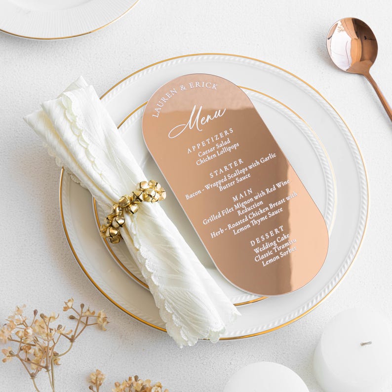 May include: A rose gold wedding menu card with rounded edges, featuring the names "LAUREN & ERICK" and a list of menu items. The card rests on a white plate with a gold rim, alongside a folded white napkin and a gold napkin ring.