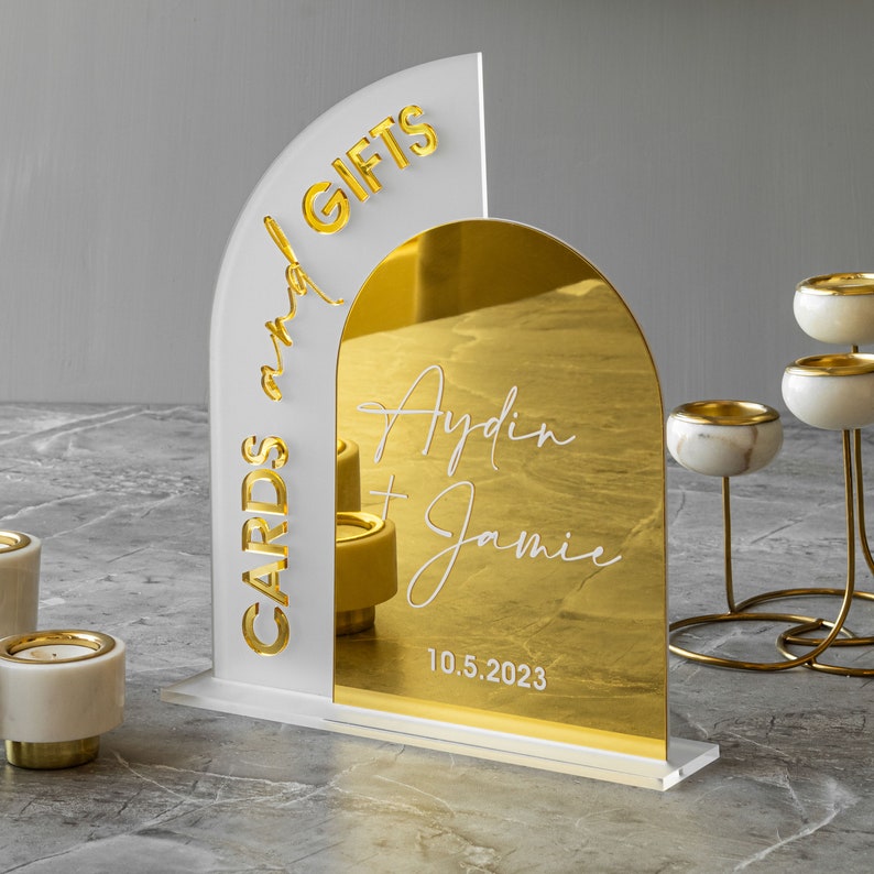 May include: White acrylic card and gift holder with a gold mirror arch. The arch has the names "Aydin + Jamie" and the date "10.5.2023" written in white script.