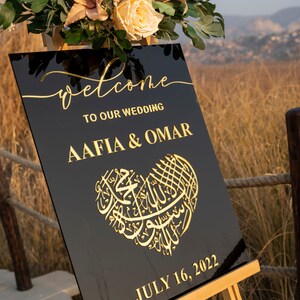 May include: Black welcome sign with gold lettering for a wedding. The sign says "Welcome to our wedding Aafia & Omar" and "July 16, 2022". The sign is decorated with a gold heart with Arabic calligraphy.