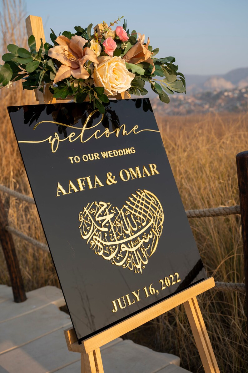 May include: Black welcome sign with gold lettering for a wedding. The sign says "Welcome to our wedding Aafia & Omar" and "July 16, 2022". The sign is decorated with a gold heart with Arabic calligraphy.