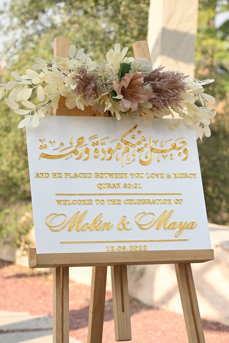 Puede incluir: Un cartel blanco con letras doradas en un caballete. El cartel dice "And he placed between you love & mercy, Quran 30:21, Welcome to the celebration of Mobin & Maya, 12.05.2022".