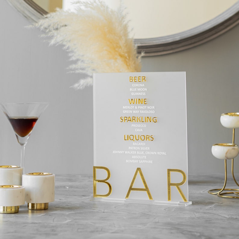 May include: A clear acrylic sign with gold lettering that reads "BEER", "WINE", "SPARKLING", "LIQUORS", and "BAR". The sign lists various beer, wine, sparkling wine, and liquor brands.