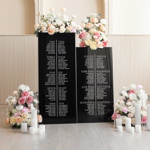 May include: Black acrylic wedding seating chart with white text listing guest names and table numbers from one to eighteen. The chart is displayed on a table with white candles and floral arrangements.