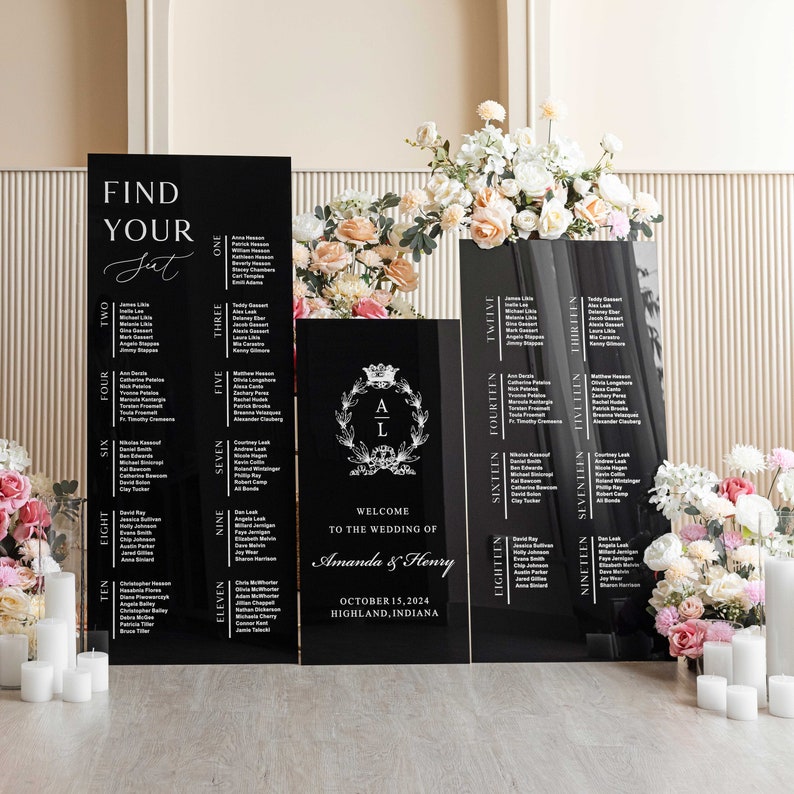 May include: Black acrylic wedding seating chart signs with white text. The signs are arranged in a row on a table with white candles. The signs include the names of guests and table numbers. The center sign features a wreath with the letters A and L in the center. The sign also includes the text "Welcome to the wedding of Amanda & Henry" and the date and location of the wedding.