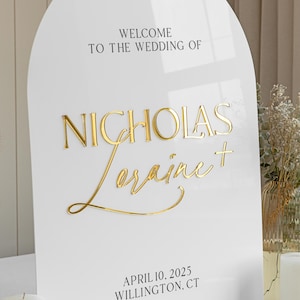 Puede incluir: Un letrero de boda de acr&iacute;lico blanco en forma de arco con letras doradas en 3D. El texto dice WELCOME TO THE WEDDING OF, NICHOLAS, Loraine, y APRIL 10, 2025 WILLINGTON, CT. Los nombres est&aacute;n en una combinaci&oacute;n de fuentes serif y cursiva.