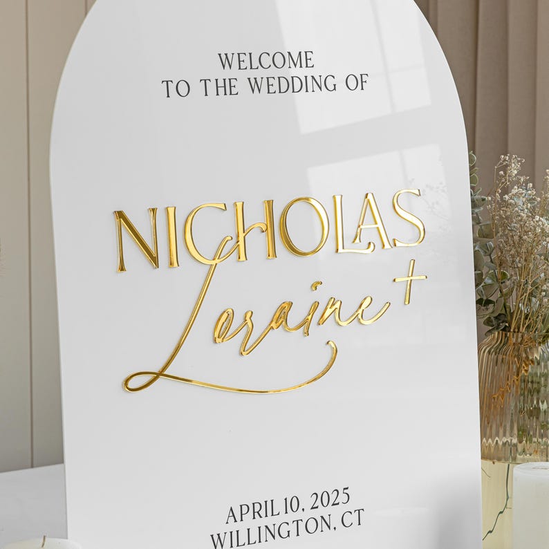 Puede incluir: Un letrero de boda de acr&iacute;lico blanco en forma de arco con letras doradas en 3D. El texto dice WELCOME TO THE WEDDING OF, NICHOLAS, Loraine, y APRIL 10, 2025 WILLINGTON, CT. Los nombres est&aacute;n en una combinaci&oacute;n de fuentes serif y cursiva.