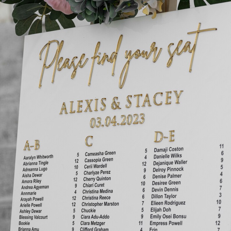 May include: A white seating chart with gold lettering for a wedding reception. The chart is titled "Please find your seat" and lists the names of guests under the headings "A-B", "C", and "D-E". The wedding date is listed as "03.04.2023" and the couple's names are listed as "Alexis & Stacey".