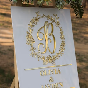 May include: White wedding sign with a gold floral wreath and the letter B in the center. The sign also includes the names Olivia & Jayden and the date 10.10.20.