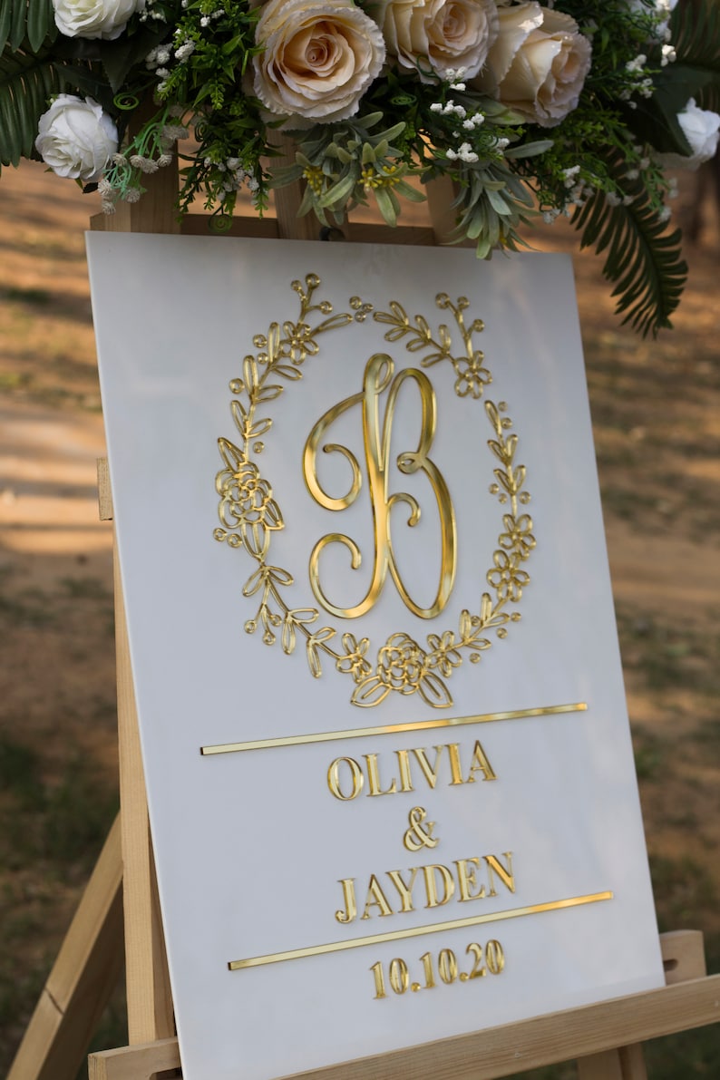 May include: White wedding sign with a gold floral wreath and the letter B in the center. The sign also includes the names Olivia & Jayden and the date 10.10.20.