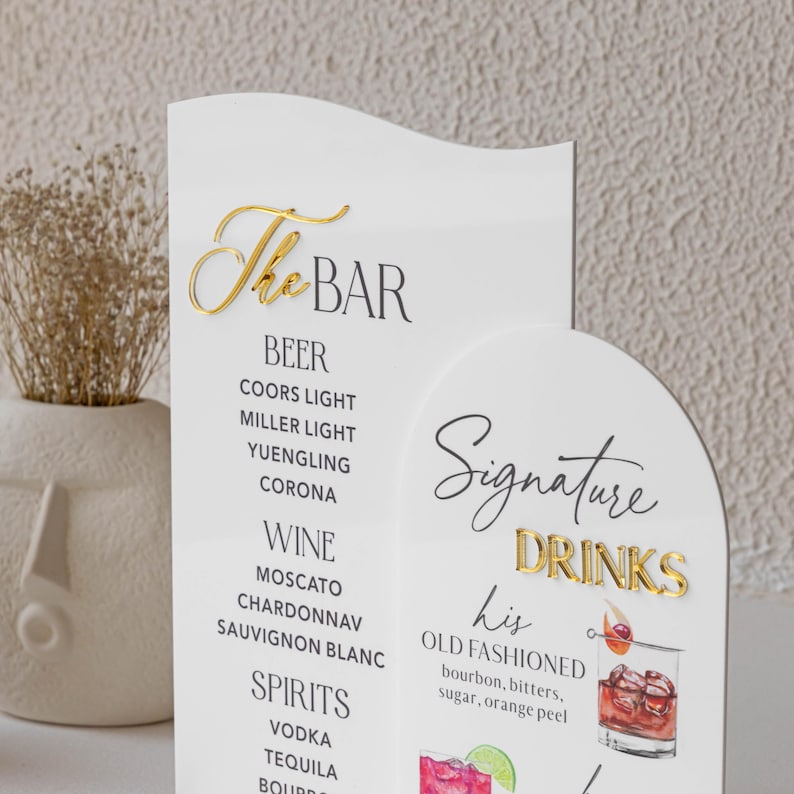 May include: A white bar menu with gold lettering that reads "The BAR" and "Signature DRINKS." The menu lists beer, wine, and spirits, including an "Old Fashioned" cocktail recipe. A decorative piece for a wedding or event.