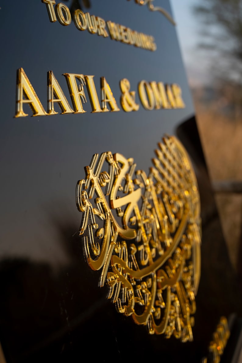 May include: A black sign with gold lettering that reads "To Our Wedding Aafia & Omar". The sign also features a gold heart-shaped design with intricate Arabic calligraphy.