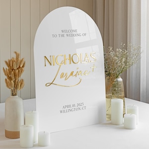 Puede incluir: Un letrero de boda de acr&iacute;lico blanco en forma de arco con letras doradas en relieve que dicen Welcome to the wedding of Nicholas + Loraine, April 10, 2025, Willington, CT. El letrero tiene un acabado liso y brillante con un borde superior redondeado.