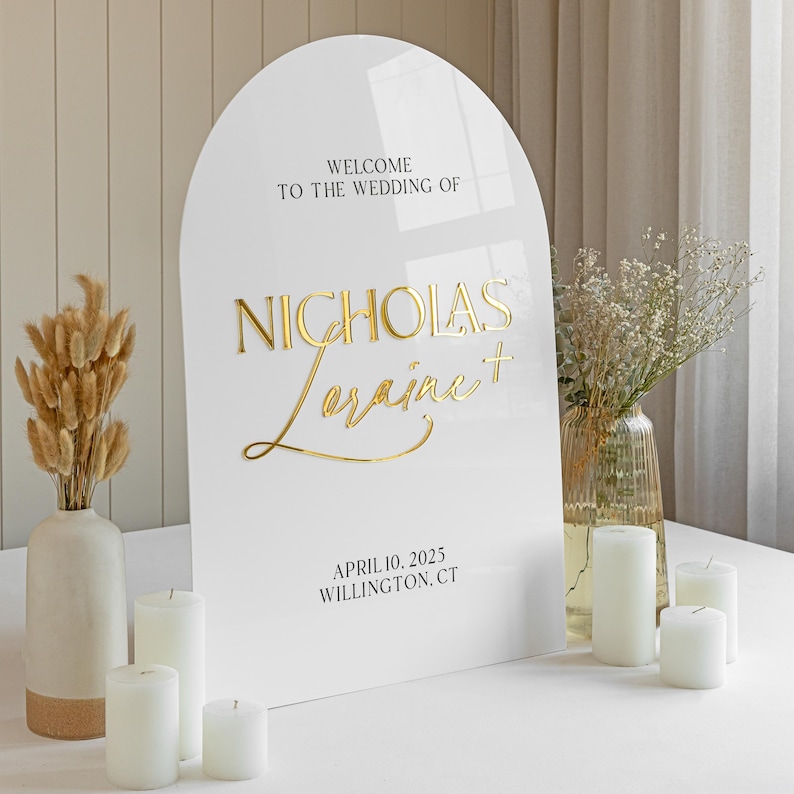 Puede incluir: Un letrero de boda de acr&iacute;lico blanco en forma de arco con letras doradas en relieve que dicen Welcome to the wedding of Nicholas + Loraine, April 10, 2025, Willington, CT. El letrero tiene un acabado liso y brillante con un borde superior redondeado.