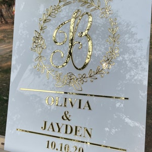 May include: White acrylic wedding sign with gold lettering. The sign features a floral wreath with the letter "B" in the center. The text below reads "Olivia & Jayden 10.10.20".