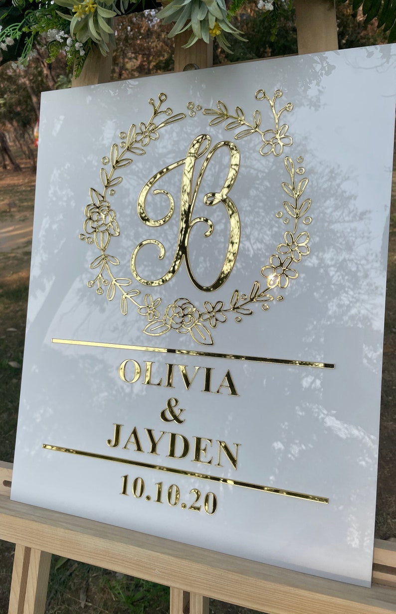 May include: White acrylic wedding sign with gold lettering. The sign features a floral wreath with the letter "B" in the center. The text below reads "Olivia & Jayden 10.10.20".