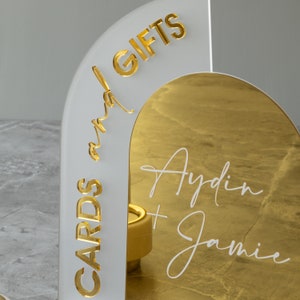 May include: White acrylic arch with gold lettering that reads "Cards and Gifts". The arch has a gold mirror backing with white lettering that reads "Aydin + Jamie".