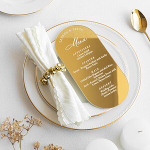 May include: A gold-colored menu card with rounded edges, featuring the names "LAUREN & ERICK" and a menu with appetizers, starter, main course, and dessert options. The menu is placed on a white plate with a gold rim, alongside a white napkin and gold napkin ring.