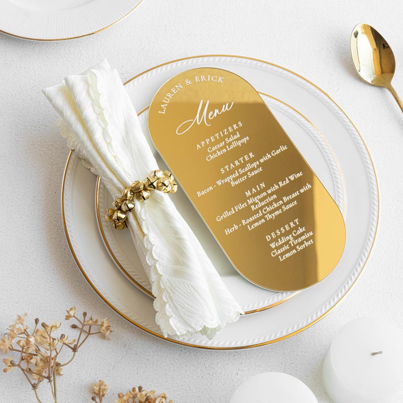 May include: A gold-colored menu card with rounded edges, featuring the names "LAUREN & ERICK" and a menu with appetizers, starter, main course, and dessert options. The menu is placed on a white plate with a gold rim, alongside a white napkin and gold napkin ring.