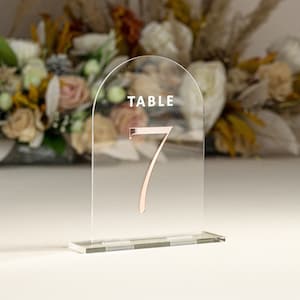 May include: Clear acrylic table number holder with a rose gold number 7 and the word "TABLE" printed on the top.