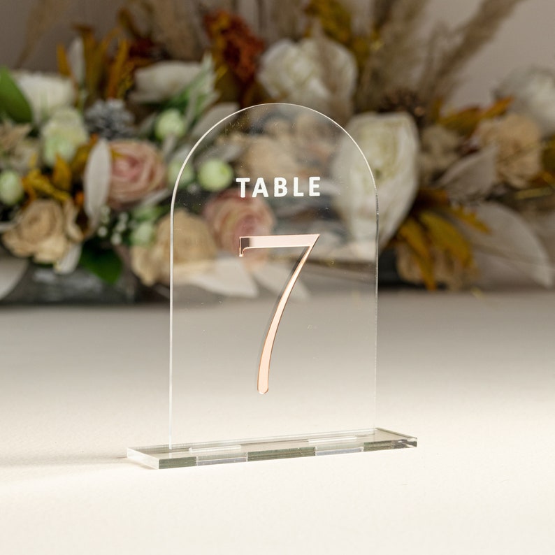 May include: Clear acrylic table number holder with a rose gold number 7 and the word "TABLE" printed on the top.