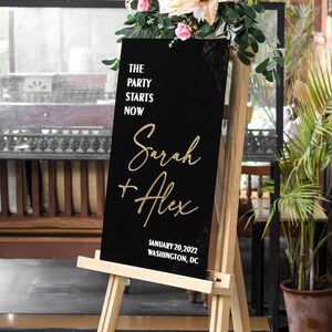 May include: Black acrylic wedding sign with gold lettering. The sign reads "The party starts now, Sarah + Alex, January 20, 2022, Washington, DC".