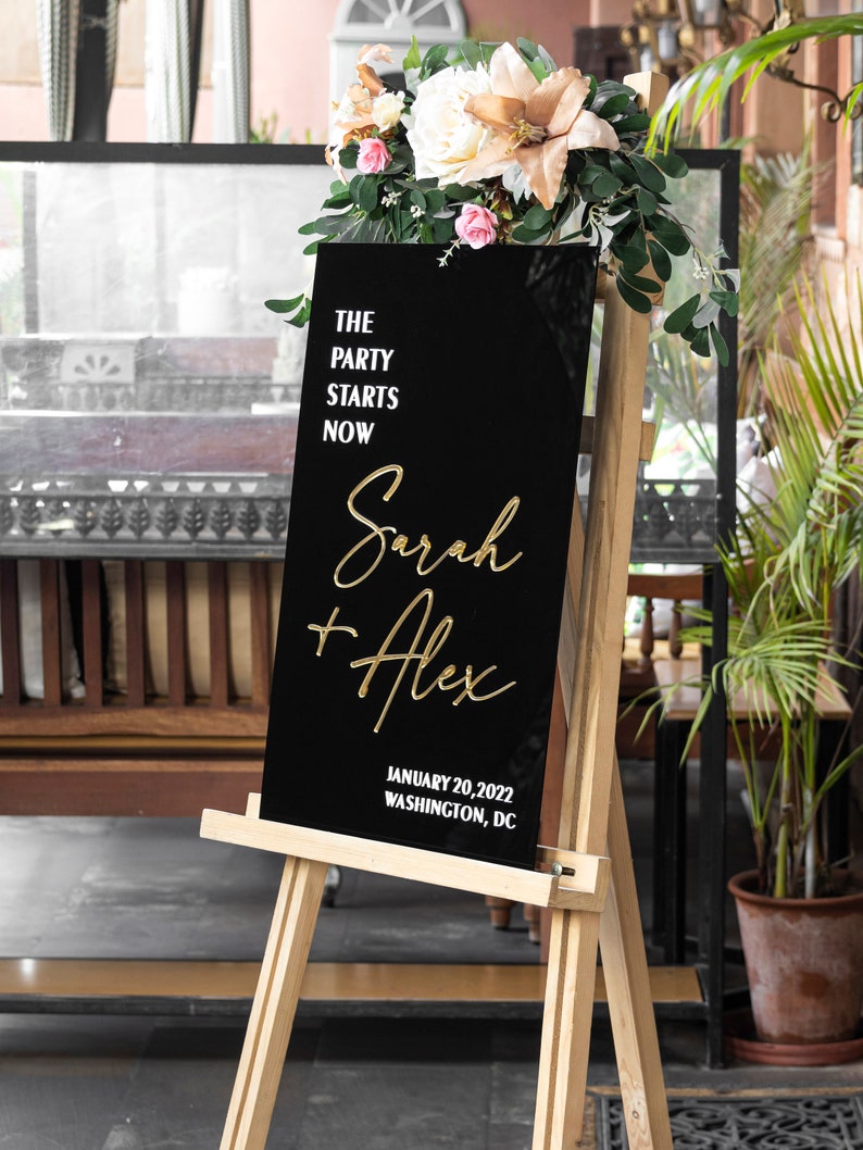 May include: Black acrylic wedding sign with gold lettering. The sign reads "The party starts now, Sarah + Alex, January 20, 2022, Washington, DC".