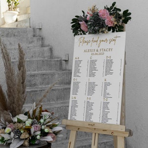 May include: A white wedding seating chart with gold lettering on an easel. The chart lists guest names alphabetically with table numbers. The text at the top reads "Please find your seat" and "Alexis & Stacey 03.04.2023".