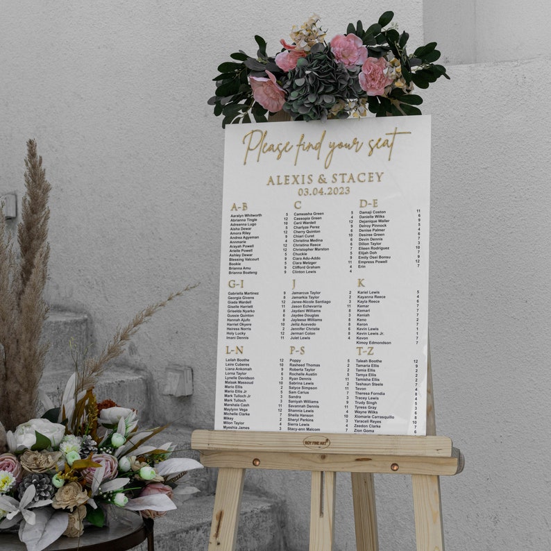 May include: A white seating chart with gold lettering on an easel. The chart lists guest names alphabetically for a wedding reception for Alexis and Stacey on March 4, 2023.