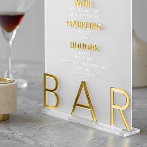 May include: A white acrylic sign with gold lettering that reads "WINE", "MERLOT & PINOT NOIR", "GREEN WAY FAVOLOSO", "SPARKLING", "PROSECCO", "CAVA", "LIQUORS", "BACARDI", "PATRON SILVER", "JOHNNY WALKER BLUE, CROWN ROYAL", "ABSOLUTE", "BOMBAY SAPPHIRE", and "BAR".