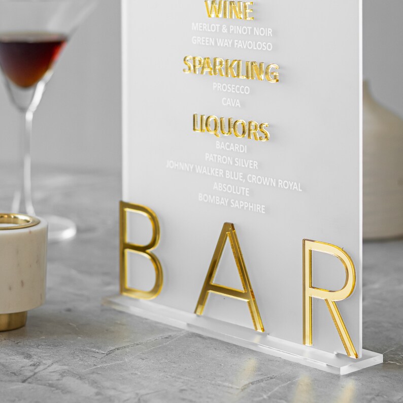 May include: A white acrylic sign with gold lettering that reads "WINE", "MERLOT & PINOT NOIR", "GREEN WAY FAVOLOSO", "SPARKLING", "PROSECCO", "CAVA", "LIQUORS", "BACARDI", "PATRON SILVER", "JOHNNY WALKER BLUE, CROWN ROYAL", "ABSOLUTE", "BOMBAY SAPPHIRE", and "BAR".