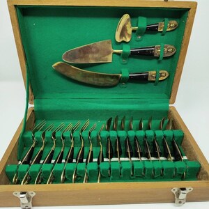 May include: A wooden box with a green velvet lining contains a set of 20 gold-toned flatware with black handles. The box also contains three decorative knives with ornate handles.