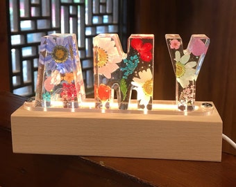 May include: A wooden base with a light switch holds four clear acrylic letters spelling "AMY". Each letter is filled with pressed flowers in shades of pink, blue, yellow, and green.