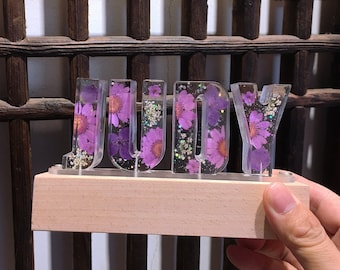 May include: A wooden base with a clear acrylic sign that spells out the name "JUDY" in all caps. The letters are filled with pressed purple flowers and glitter.