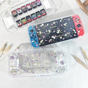 May include: Clear plastic Nintendo Switch cases with floral designs. One case holds game cartridges. Another Switch has a black background with white and pink flowers and the words "Dream" and "Explore". The third case has purple and white flowers and the words "Juliet" and "Romeo".