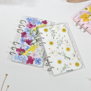 Op de afbeelding: Transparante acrylnotitieboekjes met geperste bloemen. Eén notitieboekje heeft witte madeliefjes, een ander heeft een mix van paarse, blauwe en gele bloemen. De notitieboekjes hebben zilveren ringbanden.