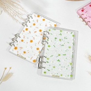 Op de afbeelding: Transparante ringband notebooks met bloemmotieven en zilveren ringbanden. Eén notebook heeft oranje bloemen, een andere heeft groene bloemen en een derde is roze met rode bloemen. De notebooks hebben de woorden "Elle" en "Hope" erop.
