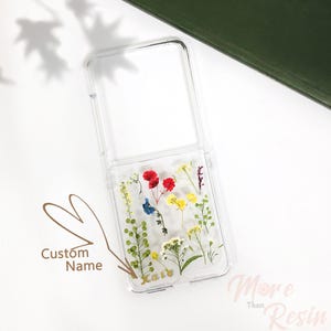 May include: A clear phone case with a floral design. The case has a clear back and a clear front with a flap that covers the screen. The flowers are pressed and preserved in resin. The case has the name "Kate" written in gold lettering on the bottom.