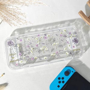 May include: Clear plastic Nintendo Switch case with embedded dried flowers and glitter. The case has the names "Juliet" and "Romeo" in gold script, and the word "Love" is also visible. A blue and black Switch controller is in the foreground.