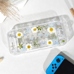 May include: Transparent gaming console case decorated with pressed daisies, blue flowers, and green leaves. The case features the words "Sun" and "New" in gold. A blue and black gaming console is also visible.