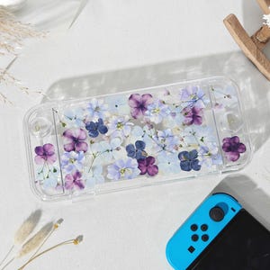 May include: A clear protective case for a gaming console, adorned with pressed purple and light blue flowers. The case is rectangular and has cutouts for buttons and ports. A blue and black game controller is visible in the image.