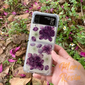May include: A clear phone case with a floral design. The case is made of two pieces that snap together. The flowers are pressed and preserved in resin. The case is designed for a Samsung Galaxy Z Flip 3.