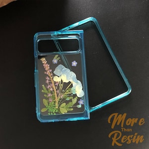 Puede incluir: Una funda de teléfono azul transparente con un diseño de flores prensadas. La funda tiene un recorte para el objetivo de la cámara y un borde elevado alrededor de los bordes para proteger la pantalla. Las flores son una mezcla de azul, morado y verde.