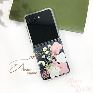 Puede incluir: Una funda de teléfono transparente con un teléfono negro en su interior. La parte posterior de la funda está llena de flores prensadas y purpurina. El nombre "Kate" está escrito en letras doradas en la funda.