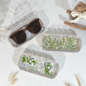 Peut inclure: Lunettes de soleil marron dans un &eacute;tui transparent. Deux &eacute;tuis transparents rectangulaires remplis de fleurs et de feuillage s&eacute;ch&eacute;s. Un &eacute;tui porte l'inscription "JUNE". Un petit cheval &agrave; bascule en bois est en arri&egrave;re-plan.