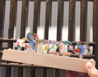 May include: A wooden base with a light wood finish holds a name spelled out in resin letters. The letters are filled with pressed flowers in shades of pink, blue, yellow, and green. The name is "Sazie" and there is a small star on either side of the name.